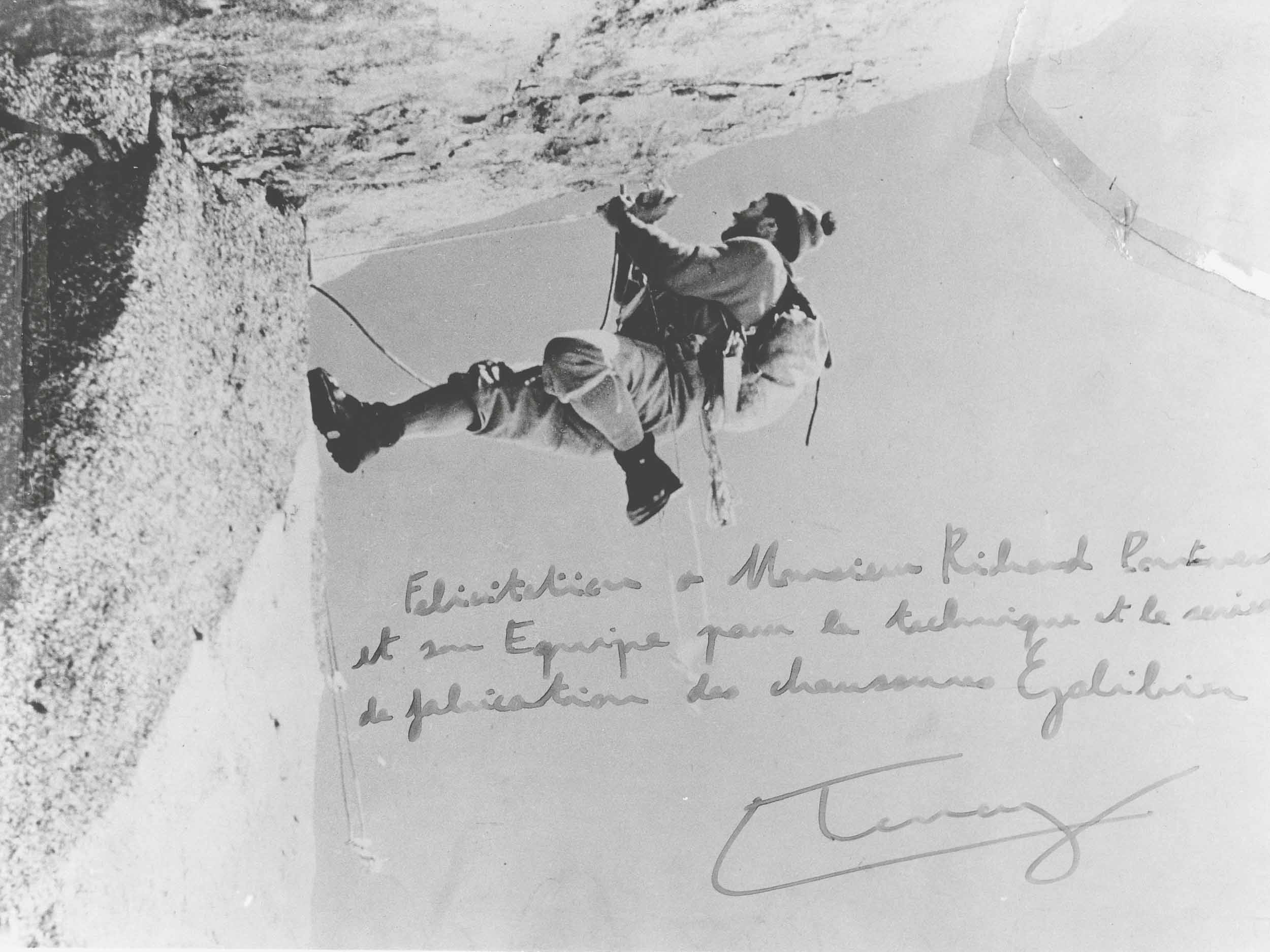Photographie d'archive de l'alpiniste Lionel Terray en plein rappel sur une paroi rocheuse. L'image comporte une dédicace manuscrite remerciant Monsieur Richard-Pontvert et son équipe pour la technique de fabrication des chaussures Galibier.