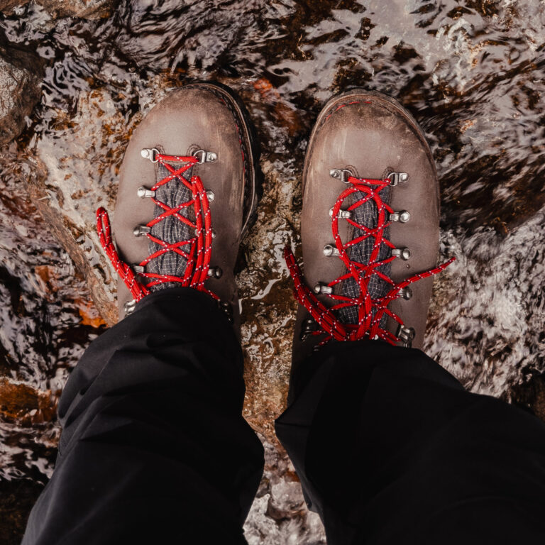 Vue de dessus de chaussures Galibier en cuir nubuck marron traversant un cours d'eau peu profond, lacets rouges contrastants.