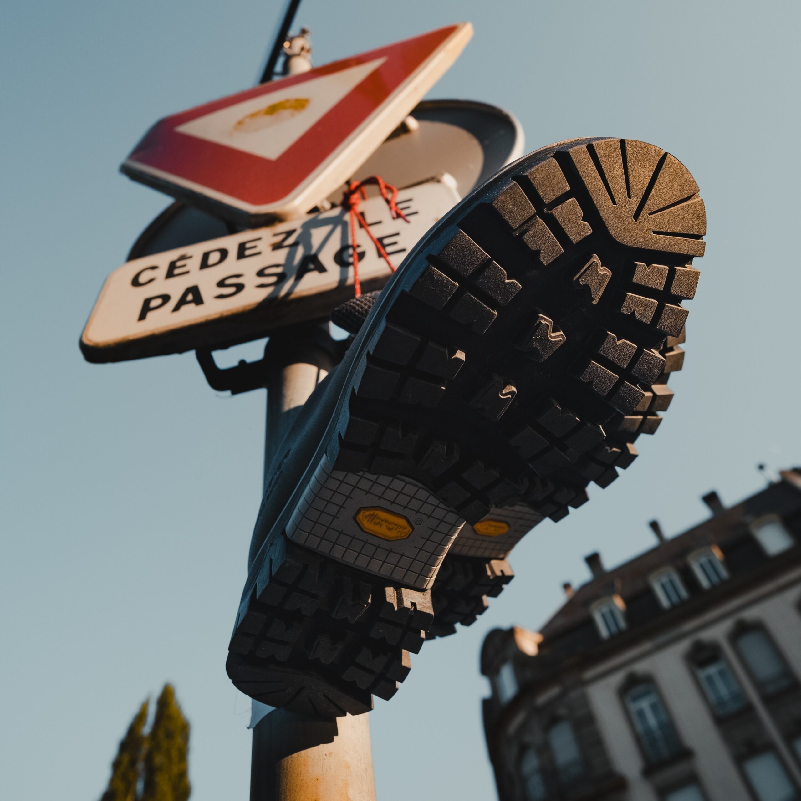 Gros plan sur une chaussure de marche urbaine Galibier Super Rando Low en cuir plein fleur, accroché via les lacets sur un panneau "Cédez le passage" en milieu urbain.