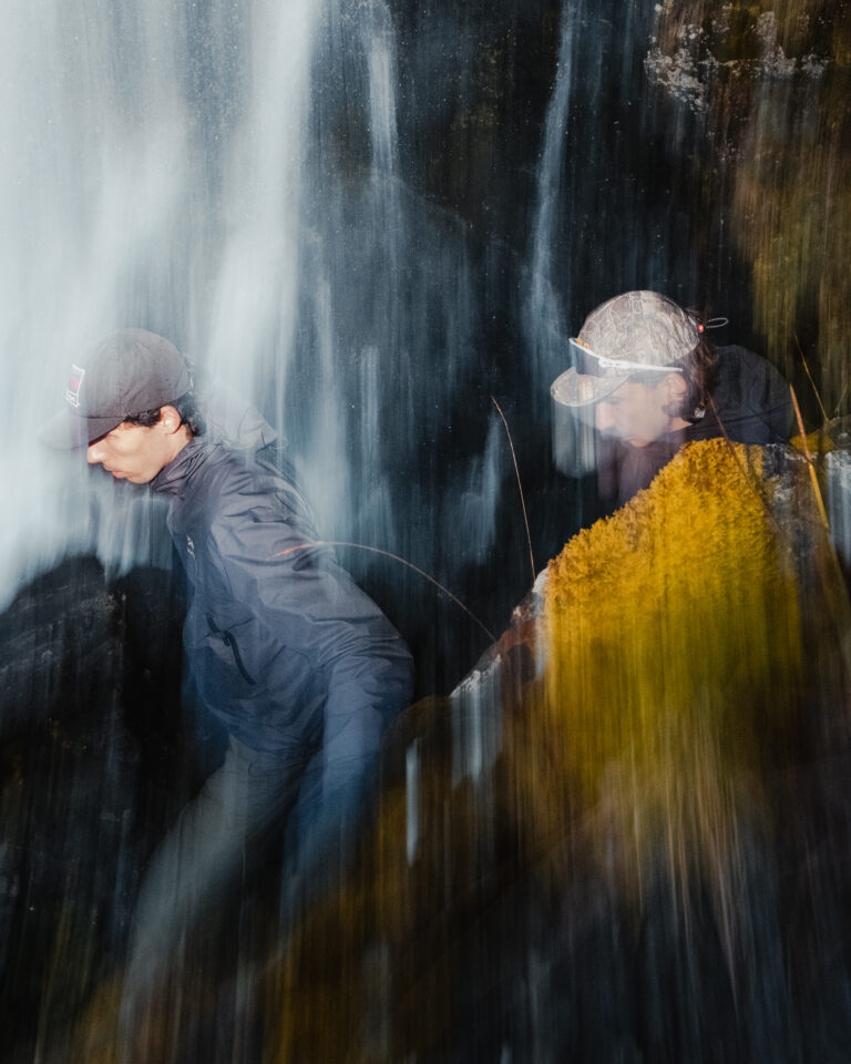 Deux randonneurs en veste technique progressent rapidement sous une cascade en montagne lors d'un speed hiking.