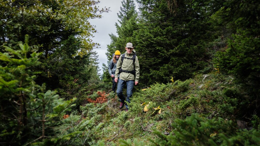 Deux randonneurs descendent un sentier technique escarpé au milieu d'une végétation de montagne dense.