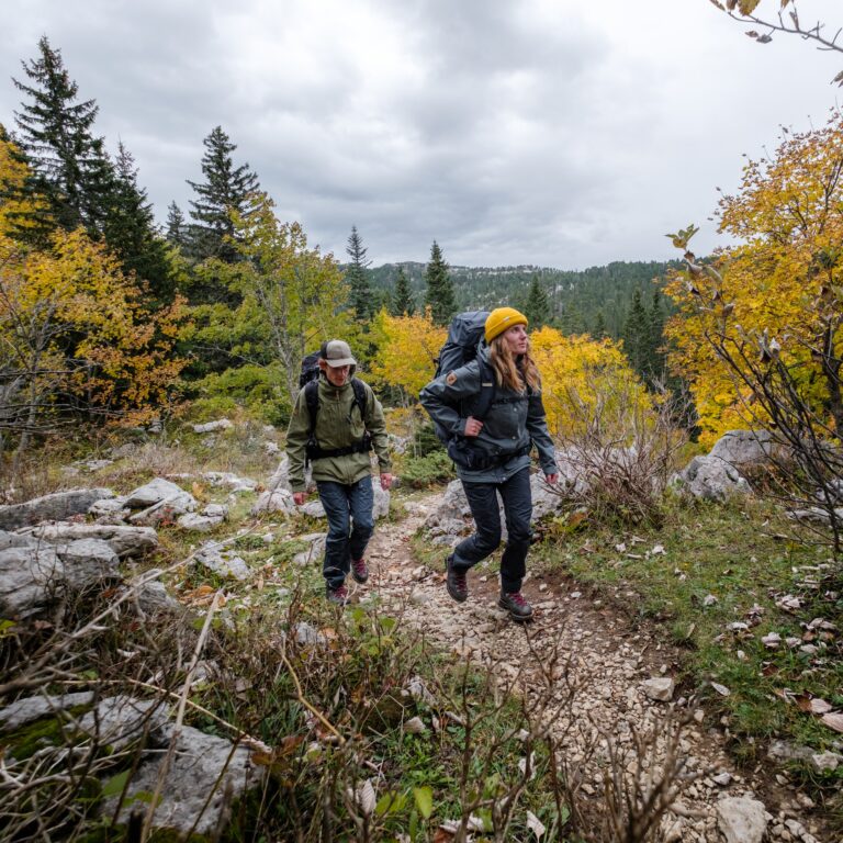 Un homme et une femme avec sacs à dos et bâtons de marche remontent un sentier escarpé dans un paysage d'automne.