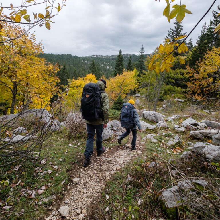 Deux randonneurs équipés de sacs à dos d'autonomie cheminent sur un sentier pierreux en sous-bois à l'automne.