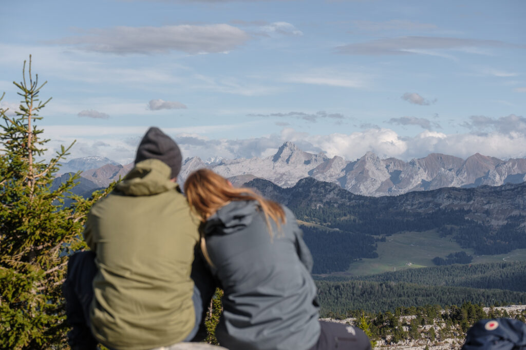 Vue de dos d'un couple assis au sommet, contemplant un vaste plateau montagneux et des forêts de sapins à l'horizon.