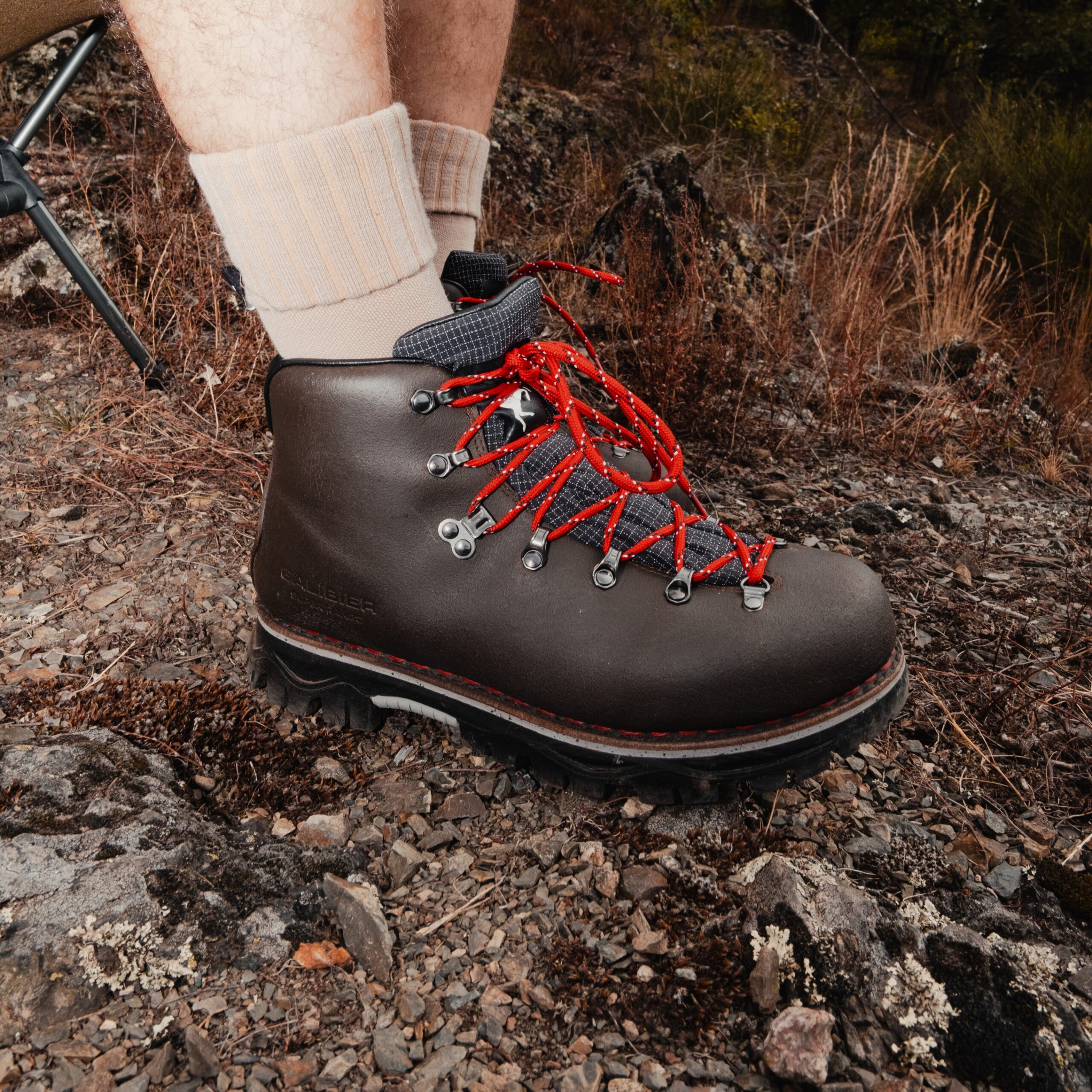 Zoom sur une chaussure de trekking Galibier en cuir marron avec lacets rouges montés sur crochets métalliques, posée sur un sol rocailleux.