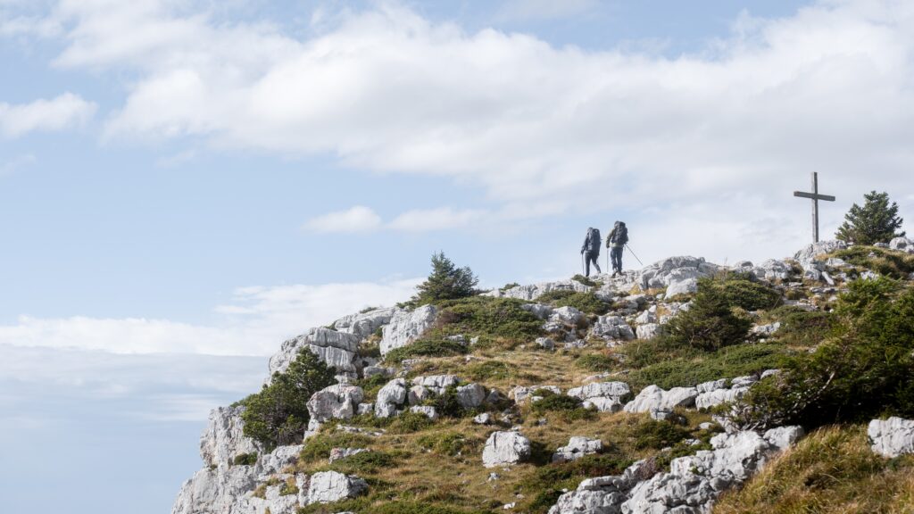 Deux randonneurs équipés de grands sacs à dos cheminent sur la crête calcaire du Parmelan, près d'une grande croix en bois.
