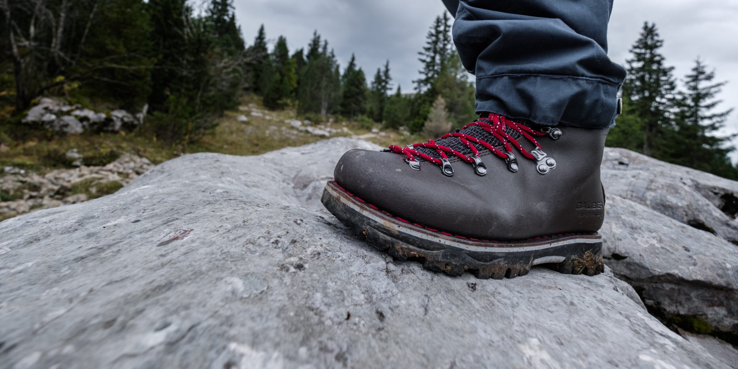 Gros plan sur une chaussure de trekking homme Galibier en cuir marron avec lacets rouges, prenant appui sur un rocher calcaire en montagne.