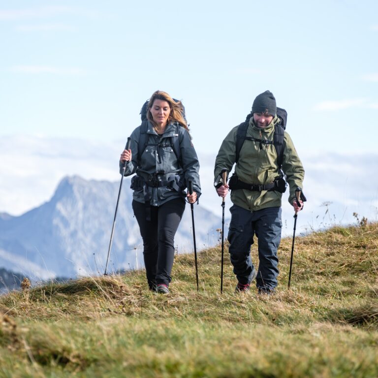 Homme et femme marchant côte à côte sur une crête herbeuse en haute montagne avec leurs bâtons et sacs de trek.