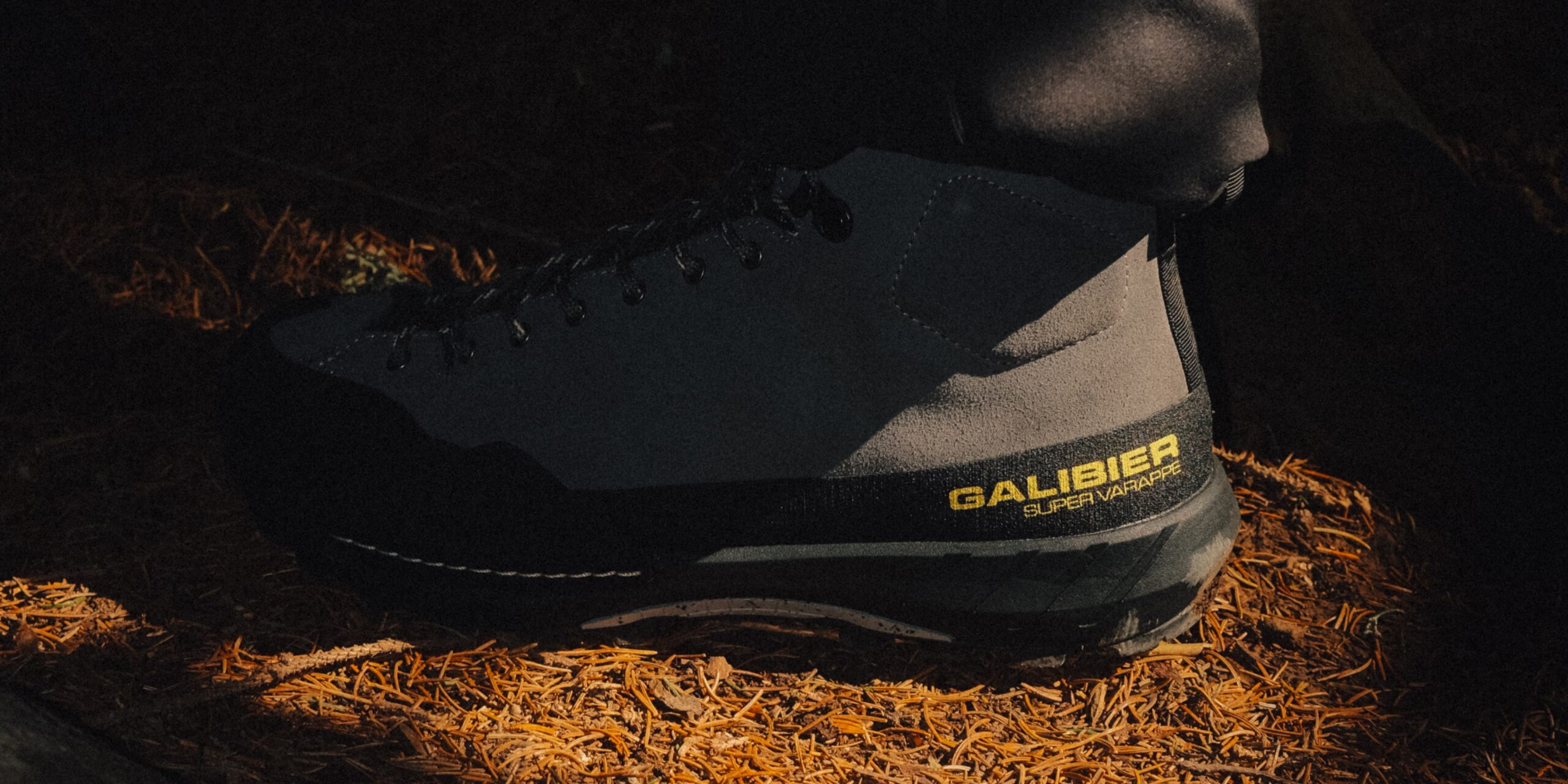 Chaussure outdoor femme Galibier Super Varappe Mid grise et noire avec logo jaune, posée sur un sol forestier couvert d'épines de pins.